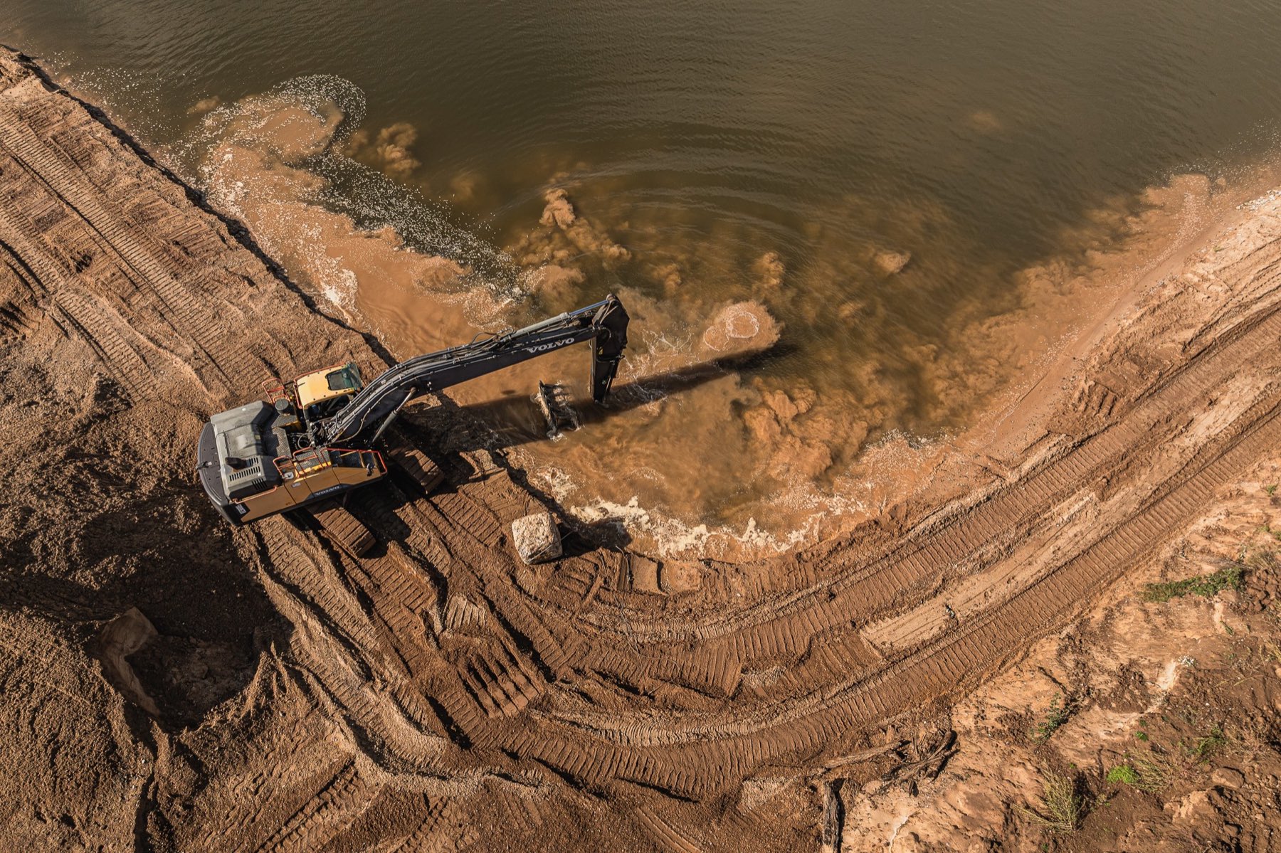 Luftbild des Nassabbaus: ein Kettenbagger arbeitet am Ufer, Sand trübt das Wasser.